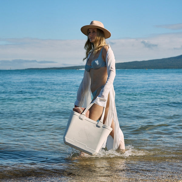 Woman walking on a beach with a large white bag, wearing a hat and light clothing.