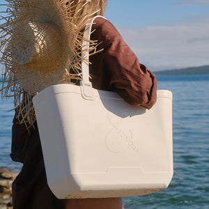 Person holding a white tote bag with a straw hat and brown jacket against a water background